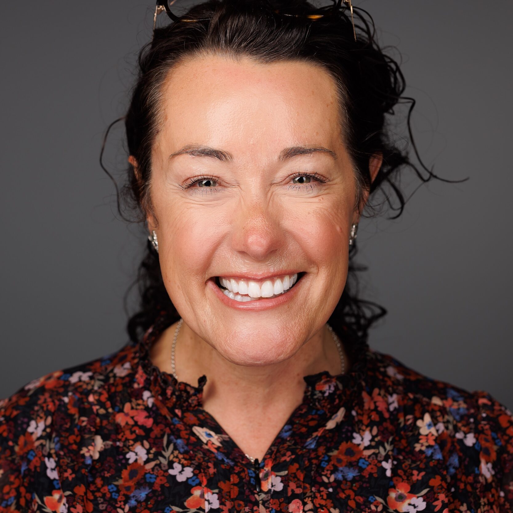 A woman with dark hair, wearing a floral blouse, smiles broadly against a plain gray background. She appears cheerful and confident, with glasses resting on her head.