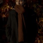 Aliyah Jay - A girl wearing a thick rust coloured scarf against a backdrop of leaves at dusk.