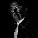 Bibi Campbell - Black and white image of an older man wearing glasses and a white shirt, dimly lit.