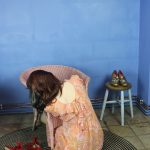 Bunny Edwards - Person in a peach dress collapsed over a rattan chair with gold heels resting on a stool against a deep blue wall.