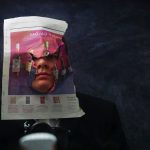 Joshua Kiddell - Portrait image of a person wearing a jacket with their head pushing through the page of a newspaper.