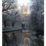 Sam Clarke - View of a cathedral from the banks of a river through frost covered leafless trees.