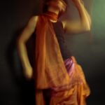 Frankie Downes - Expressive motion blurred image of a person moving with their arm above their head. They are wearing gathered burnt orange material. Rich green background