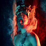 Oana Gabriela Babenco - Fantasy-like image. Portrait of a person wearing a abstract headdress with lots of different materials. Their torso naked but  material is resting over the shoulder going up and past the headdress which looks like flames. Main colours projected on the person are blue and red